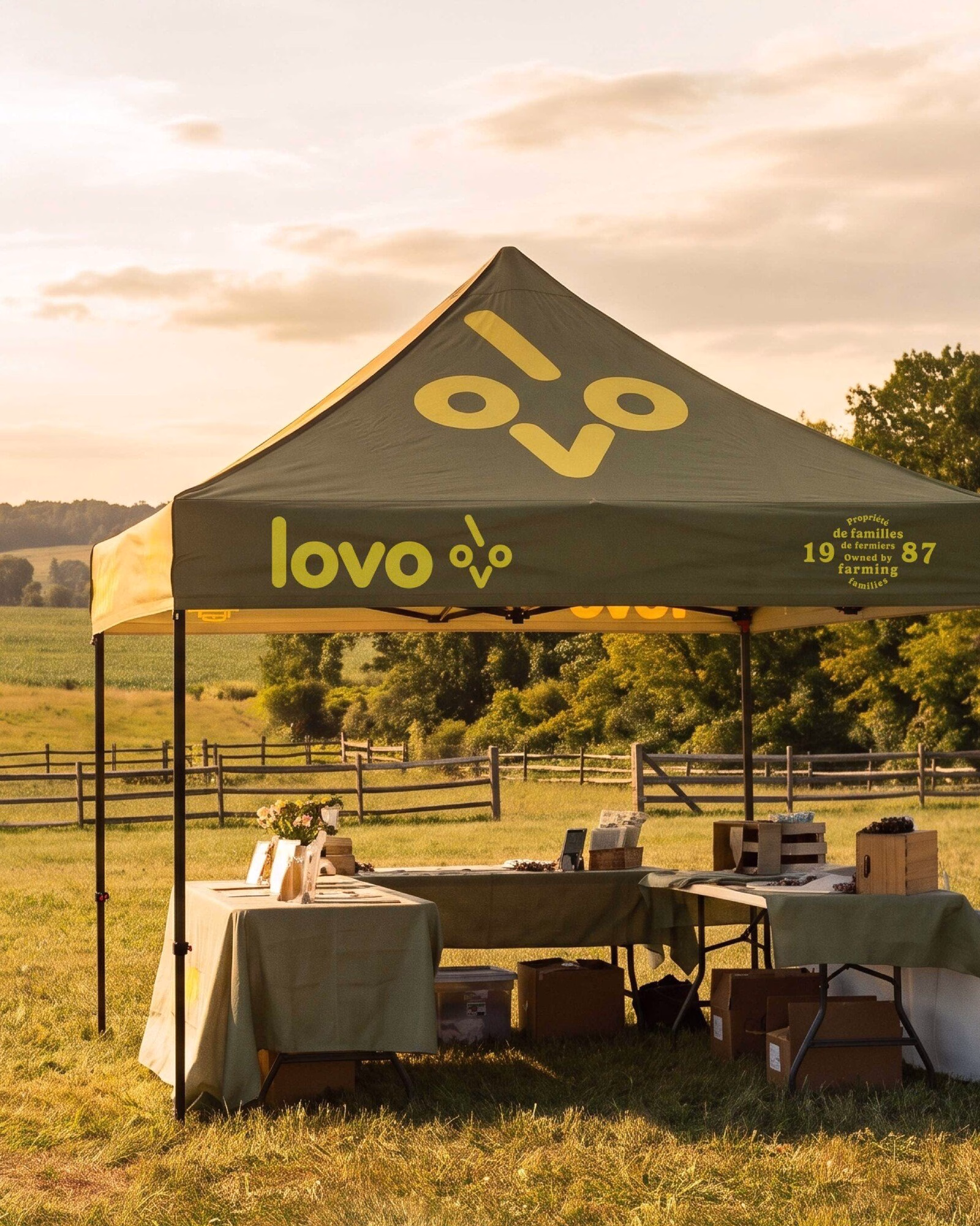 A green tent with bold yellow "lovo" branding is set up in a field, displaying farm-family products on tables.
