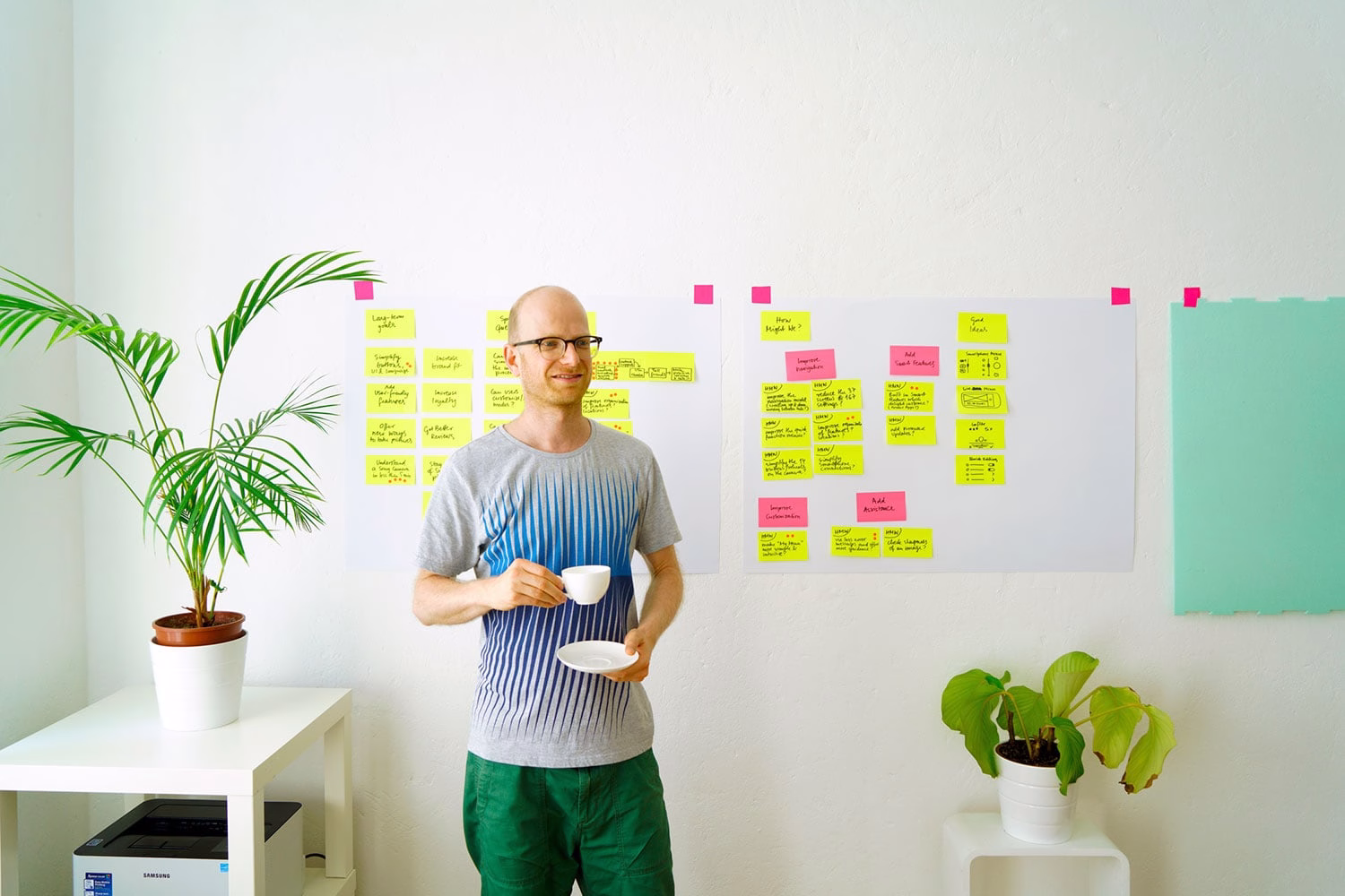 A man in a gradient blue shirt holds a teacup in a bright room with sticky notes on the wall and plants nearby.